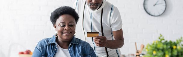 hombre afroamericano mayor sosteniendo la tarjeta de crédito cerca de esposa feliz, bandera - Foto, Imagen