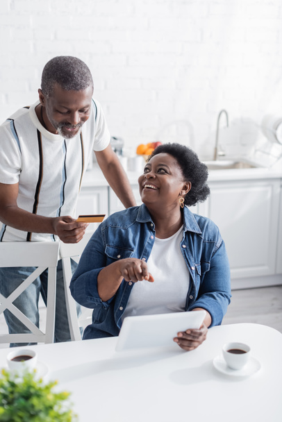 senior africano americano hombre celebración de tarjeta de crédito mientras que las compras en línea cerca feliz esposa apuntando a la tableta digital  - Foto, imagen