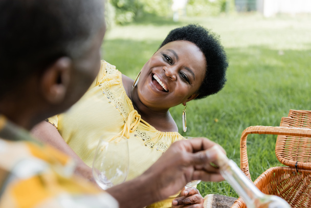 izgatott afro-amerikai nő mosolyog, és kezében borospohár közel férj piknik közben  - Fotó, kép