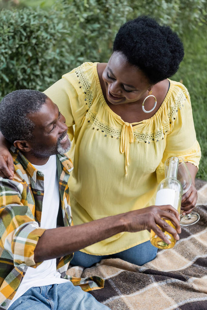 starszy Afrykański Amerykanin uśmiecha się i nalewa wino do szklanki w pobliżu szczęśliwej żony - Zdjęcie, obraz
