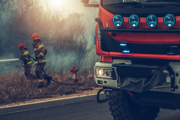 Modernes europäisches Löschfahrzeug und zwei Feuerwehrleute bekämpfen Rasenbrand im Hintergrund - Foto, Bild