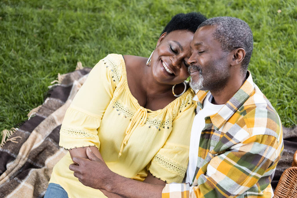 joyful and senior african american couple with closed eyes hugging during picnic  - Fotoğraf, Görsel