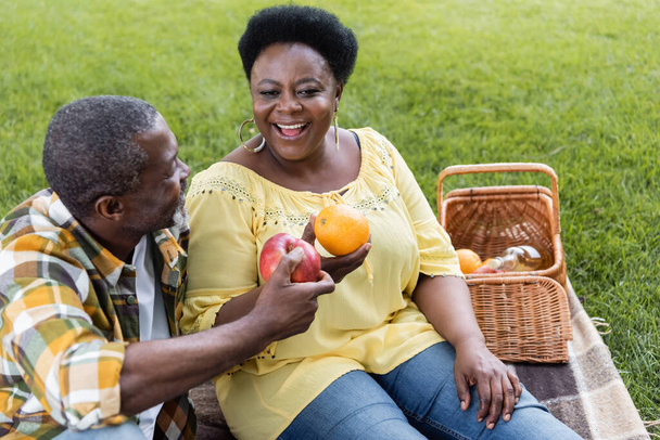 gelukkig en senior Afrikaans amerikaans paar holding fruit tijdens picknick  - Foto, afbeelding