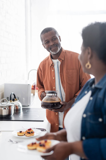 alegre sénior africano americano sosteniendo cafetera y mirando esposa - Foto, Imagen