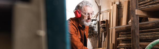 γενειοφόρος ξυλουργός με ωτοασπίδες και γυαλιά στο εργαστήριο σε θολή πρόσοψη, έμβλημα - Φωτογραφία, εικόνα
