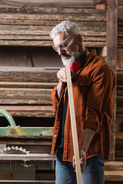 siwy projektant mebli w goglach sprawdzający drewnianą deskę w pracowni stolarskiej - Zdjęcie, obraz