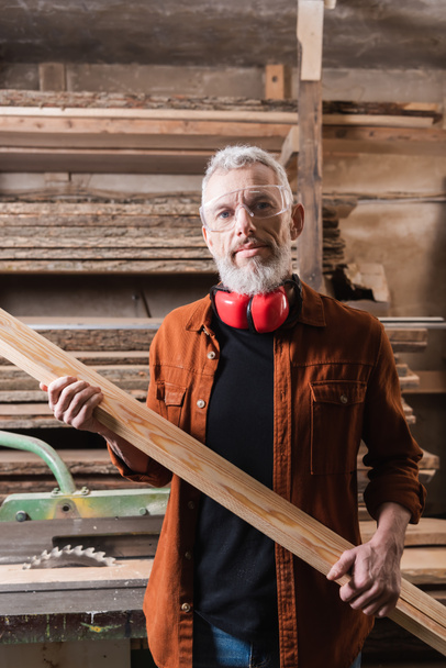 bearded woodworker with board looking at camera in workshop - Photo, Image