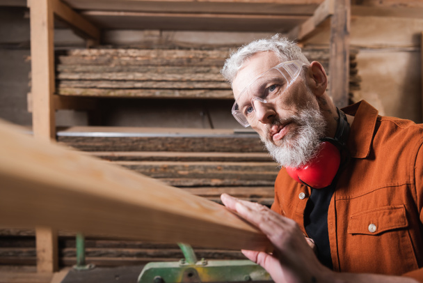 bärtiger Tischler mit Brille überprüft Holzplanke in Werkstatt - Foto, Bild
