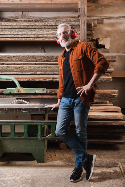 carpintero maduro de pie con la mano en la cadera cerca del banco de trabajo con sierra circular - Foto, Imagen