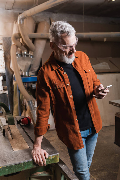 grey haired furniture designer standing with smartphone near workbench - Фото, изображение