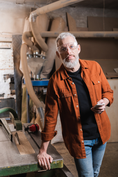 carpintero barbudo en orejeras sosteniendo smartphone y mirando a la cámara en el taller - Foto, imagen