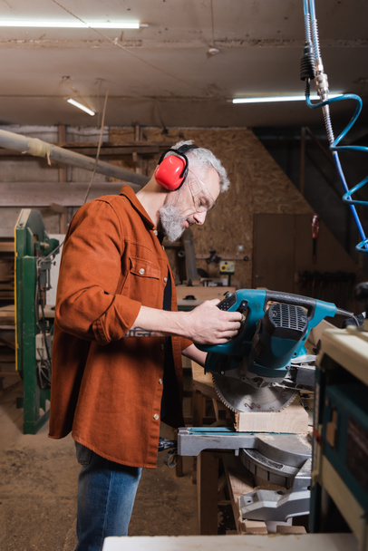 vista lateral del carpintero en orejeras protectoras cortando madera en sierra de inglete - Foto, Imagen