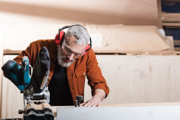 carpenter in goggles and protective earmuffs working with miter saw - Photo, Image