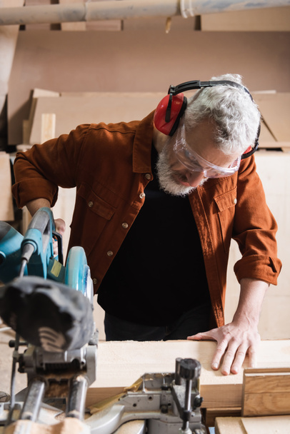grey haired woodworker cutting timber on miter saw in carpentry studio - Zdjęcie, obraz