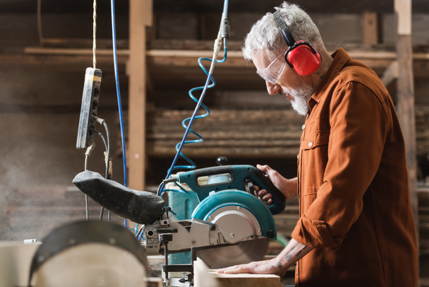 side view of bearded carpenter cutting timber on miter saw - 写真・画像