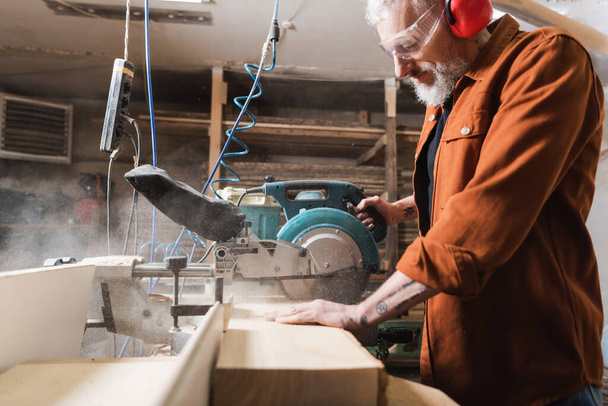 Testerenin yanındaki tahtayı kesen sakallı ahşap işçisinin yan görüntüsü - Fotoğraf, Görsel