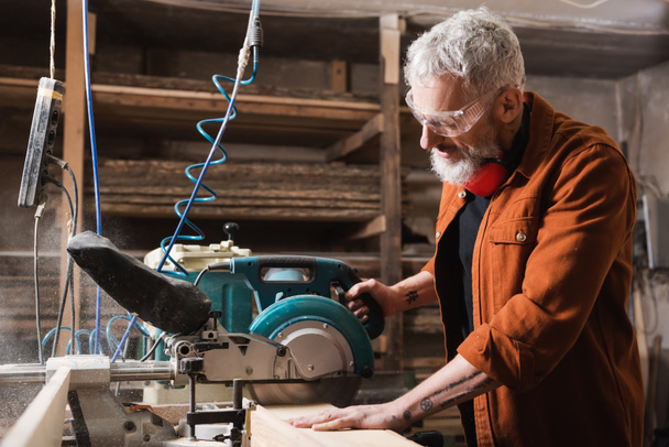 travailleur du bois mature dans des lunettes de coupe de bois avec scie à onglets - Photo, image