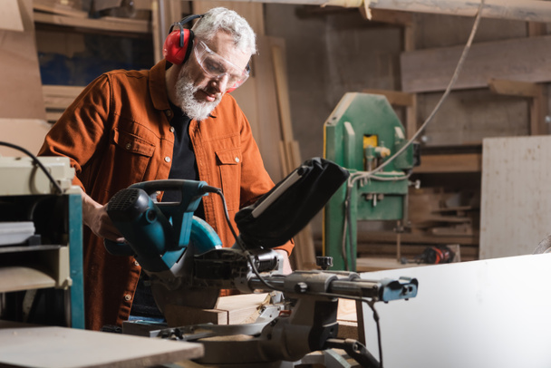 bearded joiner in goggles working on miter saw in workshop - Photo, Image