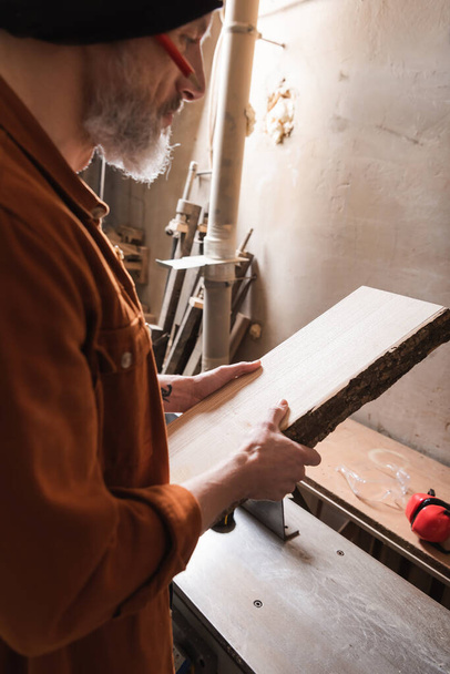 bearded woodworker in beanie holding board in workshop - Photo, Image