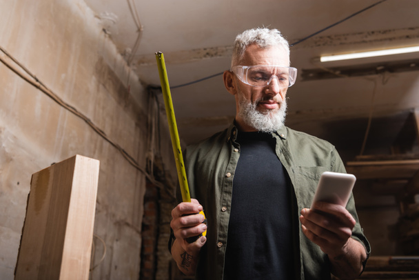 bearded carpenter in goggles holding tape measure and smartphone in workshop - Photo, Image