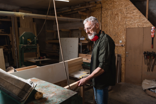Carpintero barbudo sosteniendo tablón de madera cerca de cepilladora de espesor en taller - Foto, Imagen