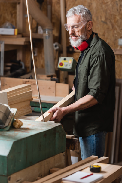 bearded carpenter holding wooden plank while working in carpentry studio - Photo, Image