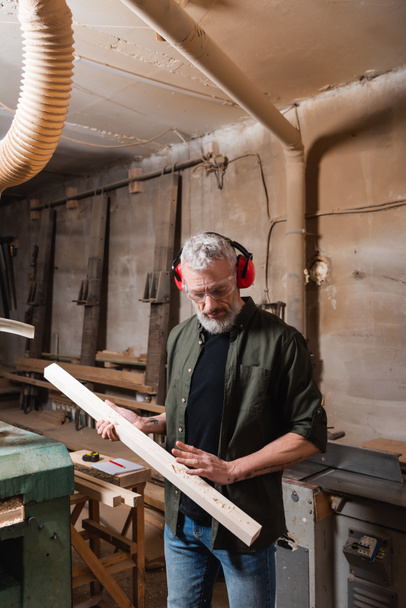 плотник в очках и наушниках проверяет деревянную доску в мастерской - Фото, изображение