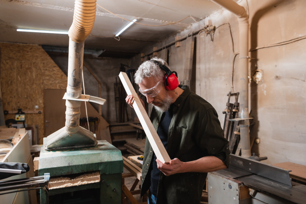 woodworker in goggles and protective earmuffs checking board in workshop - Photo, Image