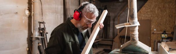carpintero en orejeras y gafas revisando tablón de madera en taller, pancarta - Foto, imagen