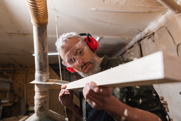 woodworker in googles and protective earmuffs checking plank in workshop - Photo, Image