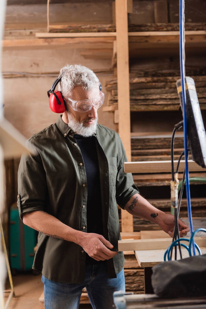 maduro carpintero mirando madera tablón en taller - Foto, Imagen