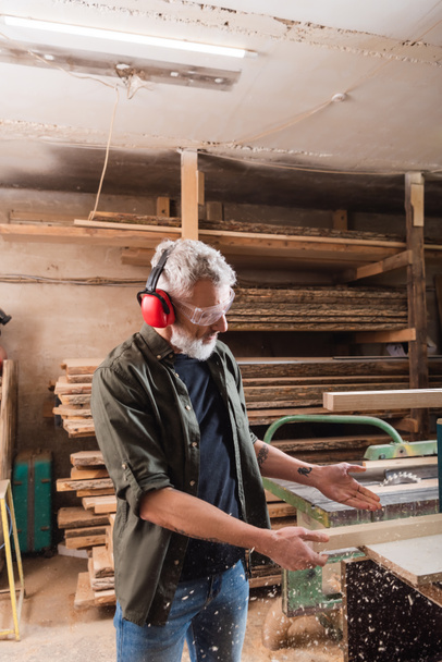 carpintero en orejeras protectoras apuntando a tablón cerca de aserrín  - Foto, Imagen