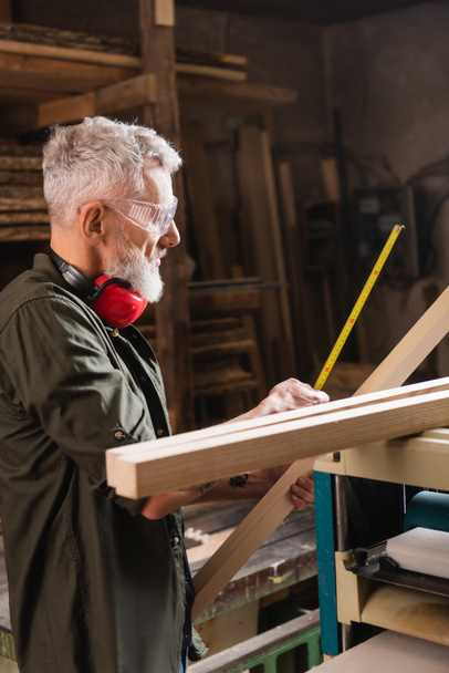 side view of bearded woodworker measuring board in workshop - Photo, Image