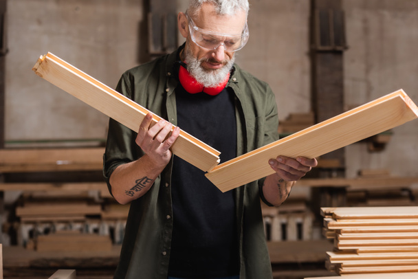 Σχεδιαστής επίπλων με τατουάζ που κρατά ξύλινες λεπτομέρειες στο στούντιο ξυλουργικής - Φωτογραφία, εικόνα