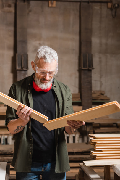 diseñador de muebles de pelo gris sosteniendo tablones de madera en taller de carpintería - Foto, Imagen
