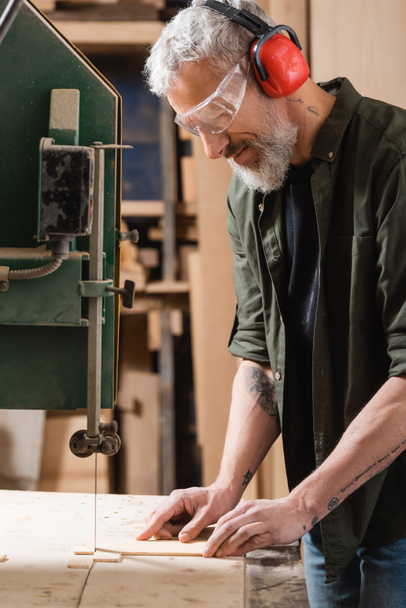 positive carpenter in goggles cutting plywood on band saw - Photo, Image