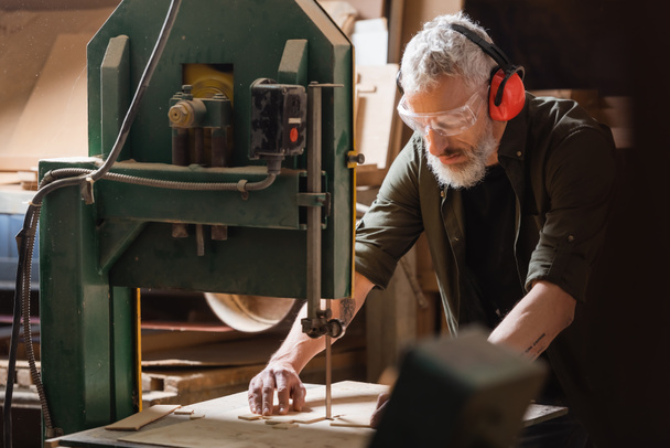 grey haired carpenter cutting plywood on band saw in workshop - Photo, Image