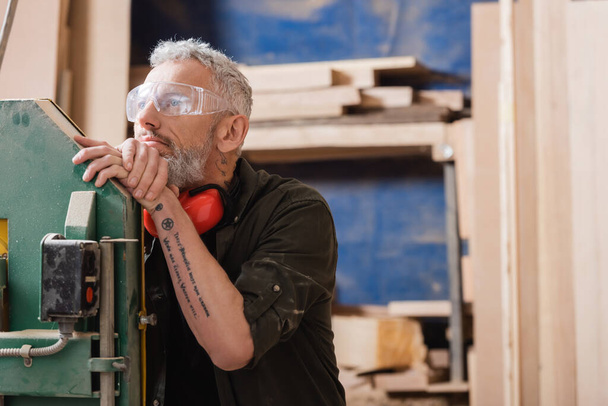 thoughtful furniture designer in goggles looking away in woodwork studio - Photo, Image