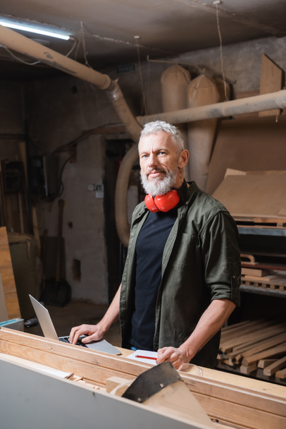 Bärtiger Möbeldesigner blickt in Holzwerkstatt in Laptopnähe auf Kamera - Foto, Bild
