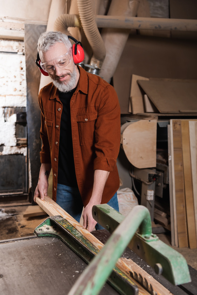 carpenter in goggles and protective earmuffs holding plank near workbench - Photo, Image