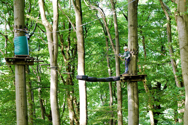 Orman macera parkında iki çocuk. Miğferli çocuklar yüksek ip üzerinde tırmanırlar. Çeviklik becerileri ve çocuklar için açık hava eğlence merkezine tırmanma. Çocuklar ve aileler için açık hava aktivitesi. - Fotoğraf, Görsel
