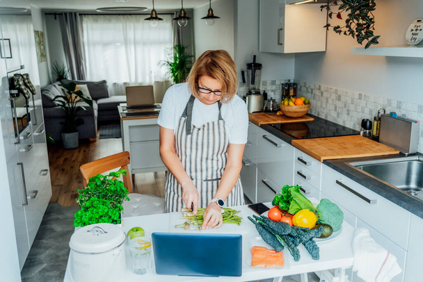 Middle age woman following a cooking tutorial video course on laptop while preparing meal in a kitchen. Woman cooking healthy dish, fish and vegetables on the table. Online recipe. Selective focus - Φωτογραφία, εικόνα