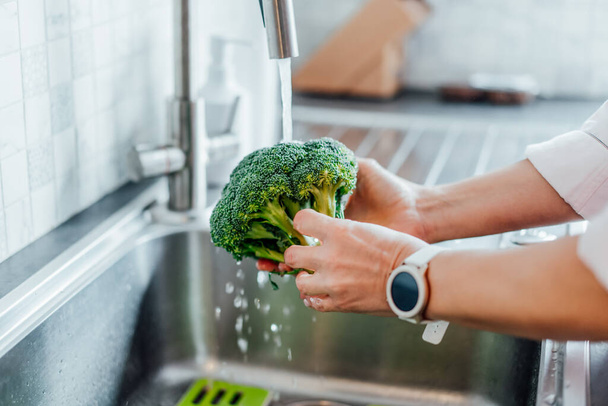 Close up woman washing fresh raw broccoli under running water over sink on her kitchen. Preparing food ingredients for lunch. Cooking healthy food. Recipes with vegetables, balanced diet. - Foto, Imagem