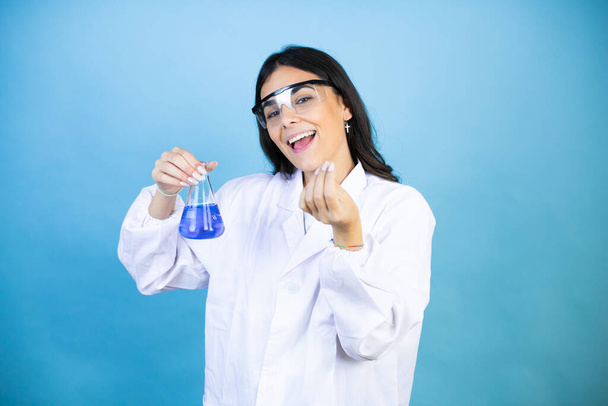 Junge brünette Frau in Wissenschaftleruniform hält Reagenzglas über isoliertem blauem Hintergrund und macht Geld-Geste mit den Händen, verlangt Gehaltszahlung, Millionärsgeschäft - Foto, Bild