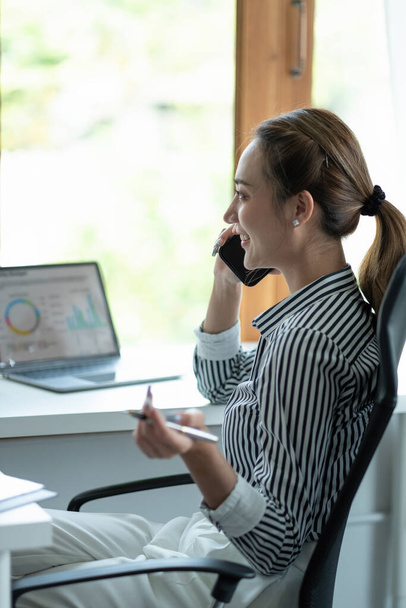 Portret Vrouwelijke Ondernemer. Vrolijke aziatische zakenvrouw praten op telefoon werken op laptop in modern kantoor. - Foto, afbeelding