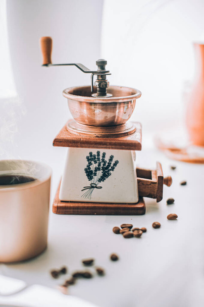 Кофемолка на белом и сером фоне с зерном кофе и чашкой горячего напитка - Фото, изображение