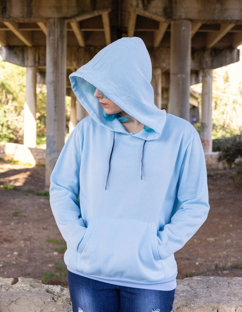 Teenager wearing light blue oversize hoodie and jeans looking aside and keeping hands in pocket. Blue haired teen girl stays outdoors against bridge pillars. Clothing mockup  - Photo, Image
