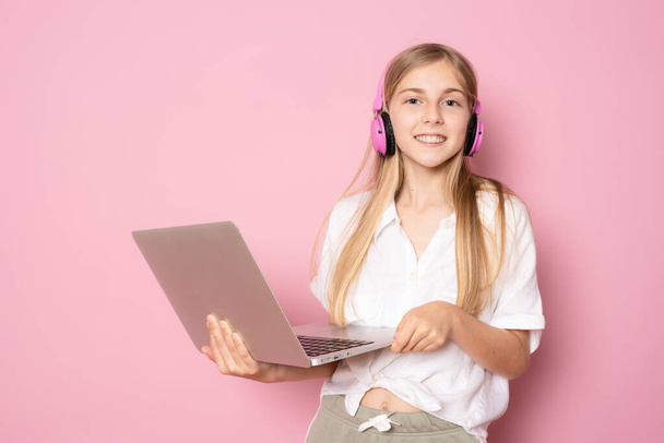 Portrait of little girl in headphones holding laptop computer while standing and looking at camera isolated over pink background - Photo, image