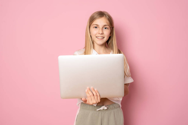 Портрет маленькой девочки с ноутбуком, стоящей и смотрящей на камеру на розовом фоне - Фото, изображение