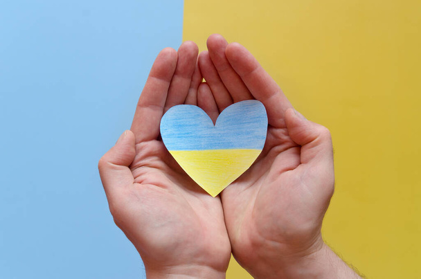 Top view of man hands holding Ukraine flag painted heart isolated on yellow, blue background. Concept symbol of help, support and no war in Ukraine - Photo, image
